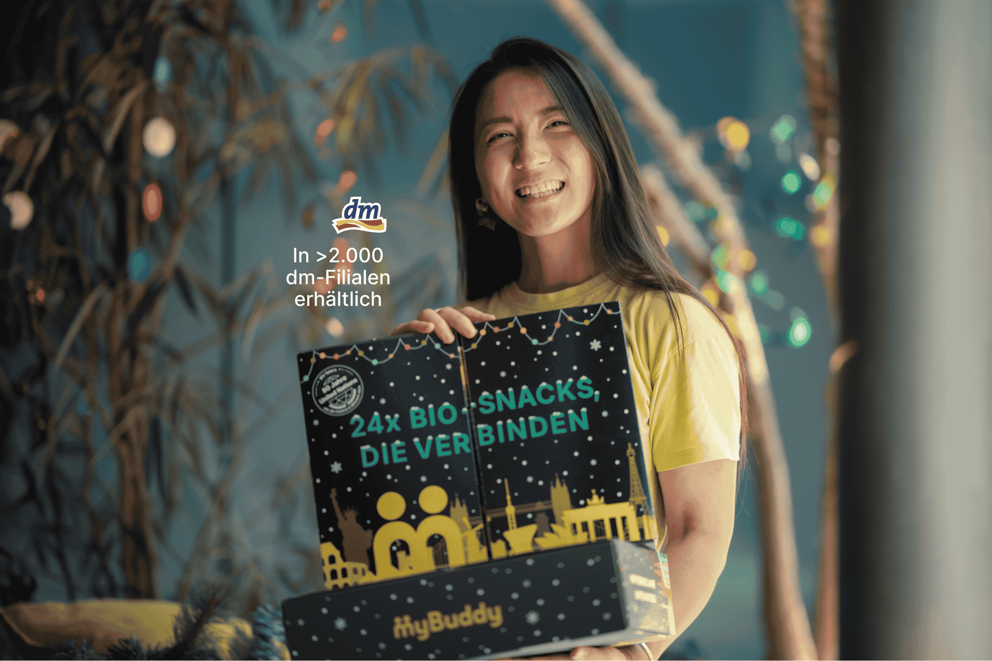 Lächelnde Frau hält den geschlossenen myBuddy Adventskalender „24x BIO-Snacks, die verbinden“ vor weihnachtlicher Deko, mit Einblendung „In über 2.000 dm-Filialen erhältlich“