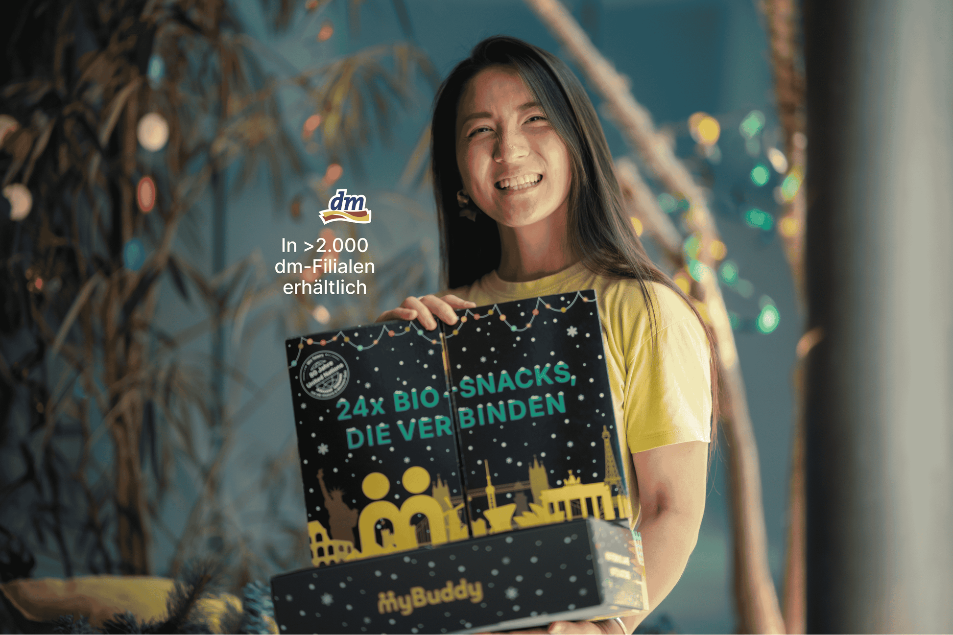 Lächelnde Frau hält den geschlossenen myBuddy Adventskalender „24x BIO-Snacks, die verbinden“ vor weihnachtlicher Deko, mit Einblendung „In über 2.000 dm-Filialen erhältlich“