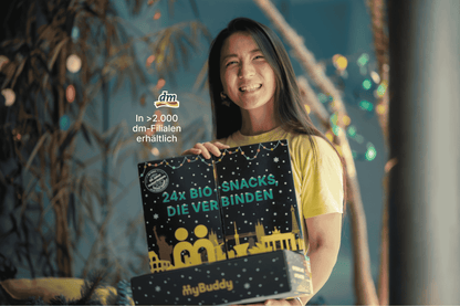 Lächelnde Frau hält den geschlossenen myBuddy Adventskalender „24x BIO-Snacks, die verbinden“ vor weihnachtlicher Deko, mit Einblendung „In über 2.000 dm-Filialen erhältlich“