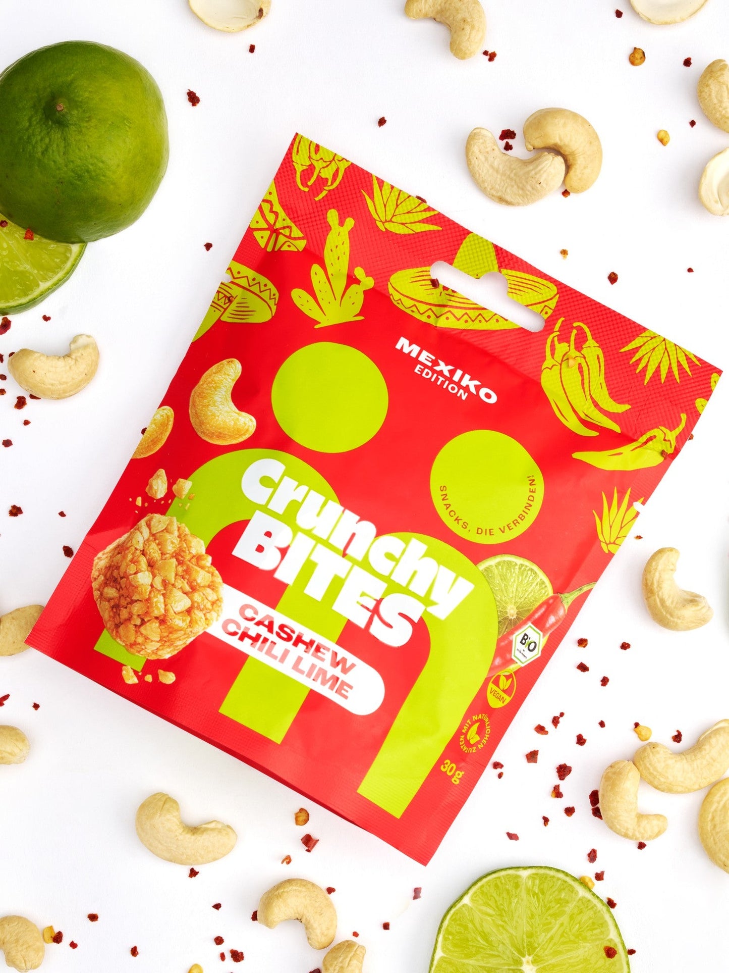 myBuddy Crunchy Nut Bites Cashew Chili Lime aus dem 9er-Set, einzelner 30-g-Beutel im Flatlay mit Limette, Cashewkernen und Chiliflocken